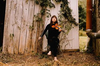 Violinistin Tessa Lark barfuss mit Ihrer Geige auf dem Arm vor einer Holzscheune