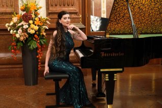 Die Pianistin sitzt in einem dunkelgr&uuml;n schimmerden Kleide an einem Fl&uuml;gel der Marike B&ouml;sendorfer. Im Hintergrund sieht der Betrachter &uuml;ppigen Blumenschmuck.