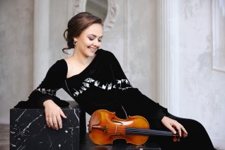 Die Geigerin lehnt sich entspannt auf eine Sitzgelegenheit mit ihrem Instrument vor hellem Hintergrund. Sie tr&auml;gt ein schwarzes festliches Kleid.