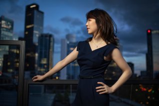 Die Pianistin steht mit Blick zur Seite am Fenster vor imposanter Gro&szlig;stadtkulisse.