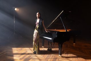 Die Pianistin st&uuml;tzt sich stehend auf dem hinter ihr stehenden Konzertfl&uuml;gel ab.