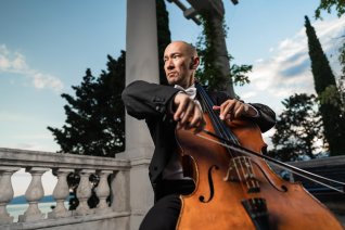 Der Cellist sitzt mit seinem Instrument auf einer Terrasse vor blauem Himmel.
